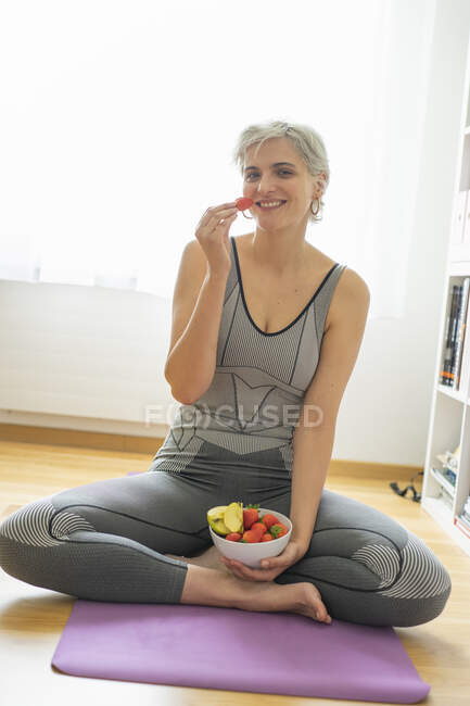 Mujer practicando yoga en casa, tomando un bocadillo de fruta saludable - foto de stock