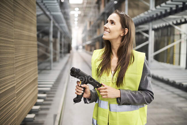 Mujer sonriente en la sala de la fábrica con chaleco de seguridad que sostiene el escáner de código de barras - foto de stock