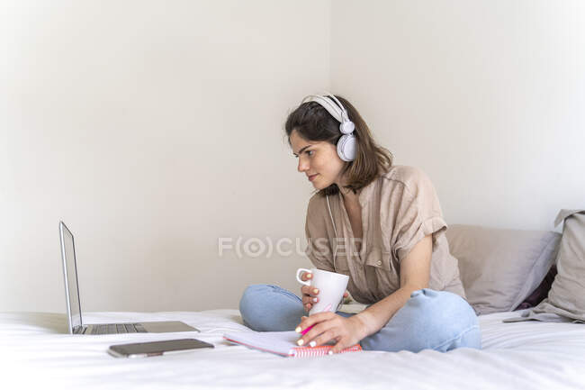 Молода жінка сидить на ліжку з навушниками і ноутбуком приймає нотатки — стокове фото
