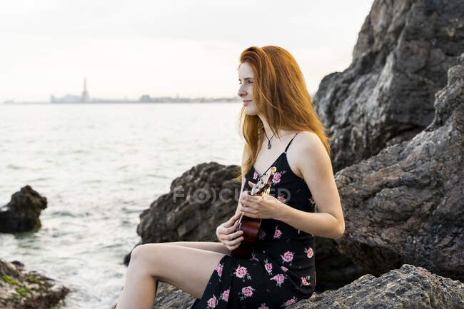 Улыбающаяся рыжая молодая женщина, сидящая на скале у моря и играющая на укулеле — стоковое фото