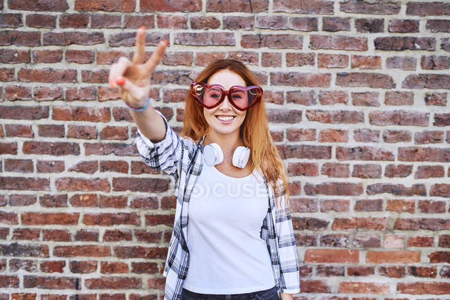 Magnifique jeune femme dans des lunettes en forme de coeur souriant à la caméra et faisant geste de paix — Photo de stock