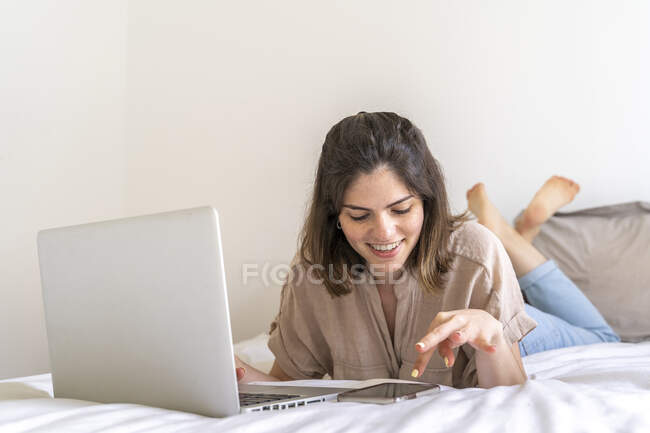 Mujer joven feliz acostada en la cama usando smartphone y portátil - foto de stock