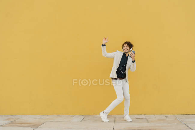Empresario cantando y bailando frente a la pared amarilla escuchando música con auriculares y smartphone - foto de stock