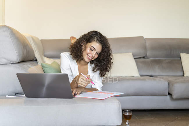 Улыбающаяся молодая женщина делает заметки и использует ноутбук на диване дома — стоковое фото