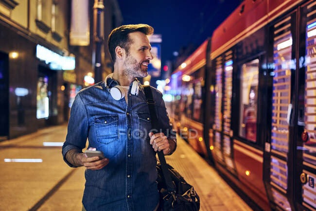 Sorrindo homem usando seu smartphone na cidade à noite enquanto espera o bonde — Fotografia de Stock