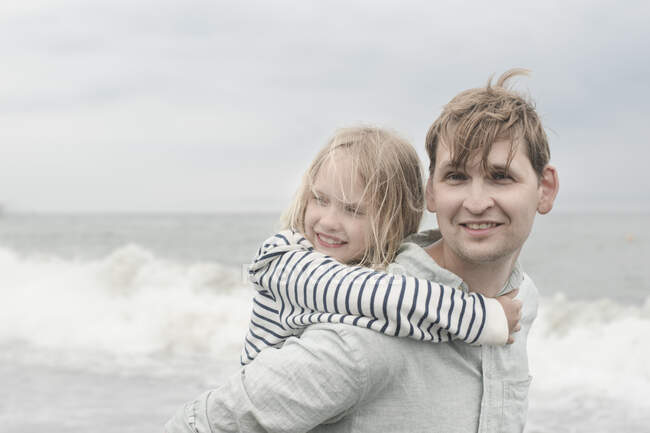 Портрет улыбающегося отца, несущего свою дочь на спине к морю — стоковое фото