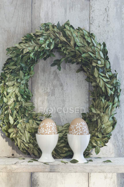 Dos huevos de Pascua pintados en tazas de huevo y corona de árbol de caja en el estante delante de la pared de madera — Stock Photo