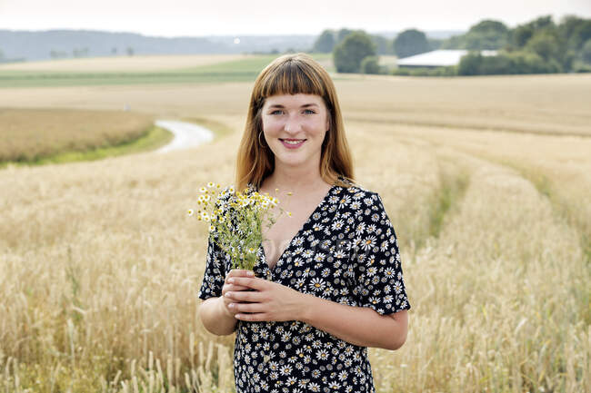 Портрет усміхненої молодої жінки з грою квітки ромашки, що стоїть перед зерновим полем — стокове фото