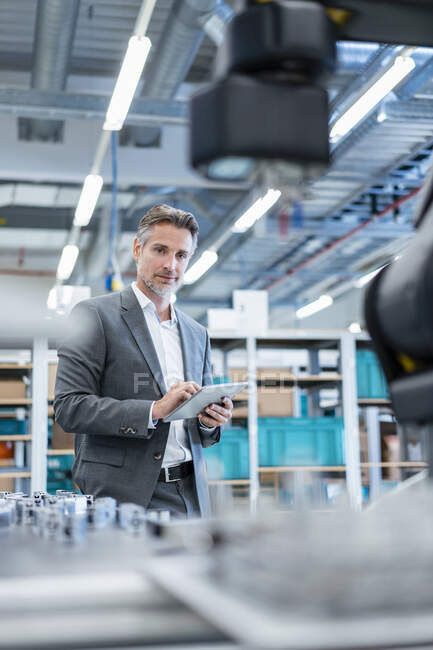 Бізнесмен з планшетом у сучасному заводському залі, який дивиться на робота — стокове фото