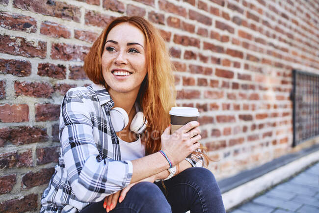 Young woman sitting against brick wall holding cup of coffee and looking away — Stock Photo
