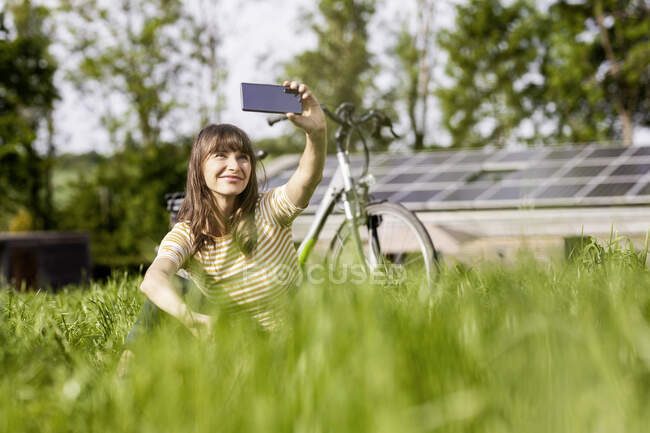 Mujer relajada sentada en un prado con bicicleta tomando una selfie - foto de stock