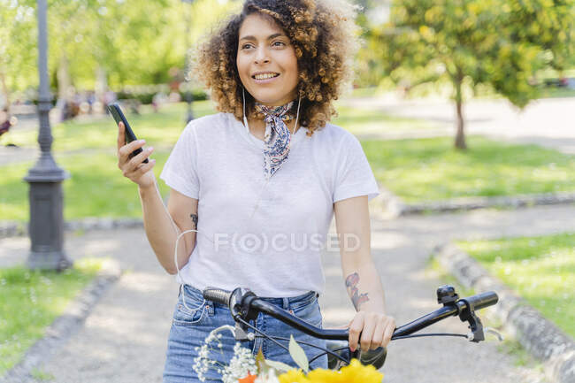 Mujer sonriente con teléfono celular, auriculares y bicicleta en el parque - foto de stock