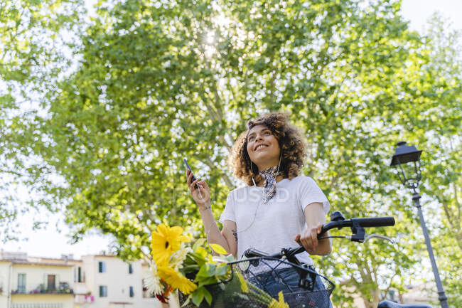 Donna sorridente con cellulare, auricolari e bicicletta nel parco — Foto stock