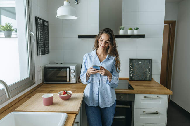 Jeune femme portant un pyjama dans la cuisine à la maison en utilisant un téléphone portable — Photo de stock