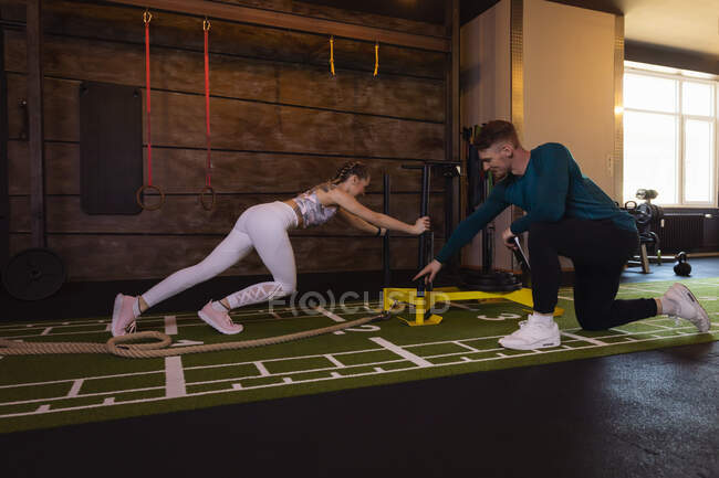 Junge Frau trainiert in Fitnessstudio mit Personal Trainer, der Gewichtschlitten schiebt — Stockfoto
