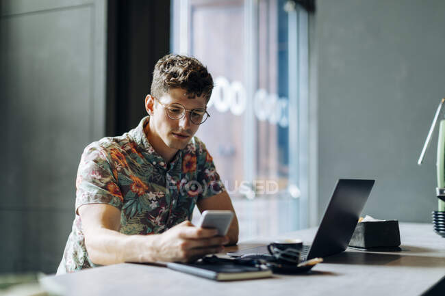 Young man sitting in cafe, using samrtphone and laptop — Stock Photo