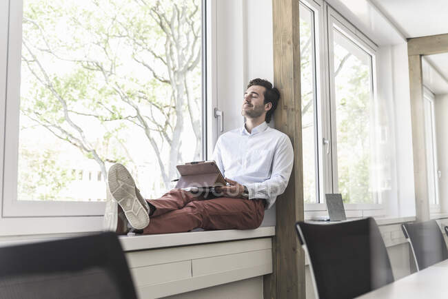 Giovane uomo d'affari con tablet digitale seduto sul davanzale della finestra, prendendo una pausa — Foto stock