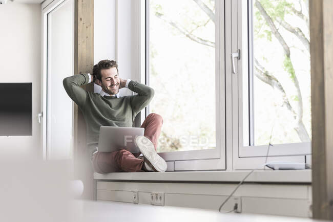 Молодий бізнесмен сидить на підвіконні, використовуючи ноутбук. — стокове фото