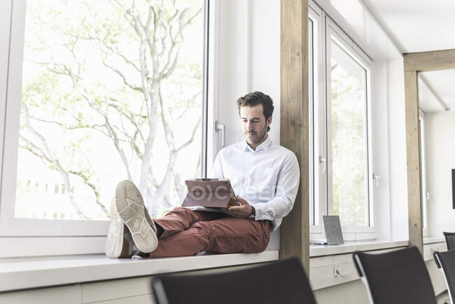 Young businessman sitting on windowsill, using digital tablet — Stock Photo
