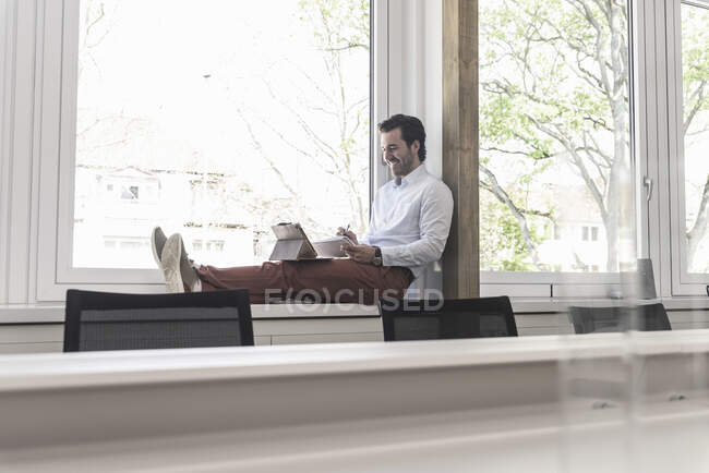 Giovane uomo d'affari seduto sul davanzale della finestra, utilizzando tablet digitale — Foto stock