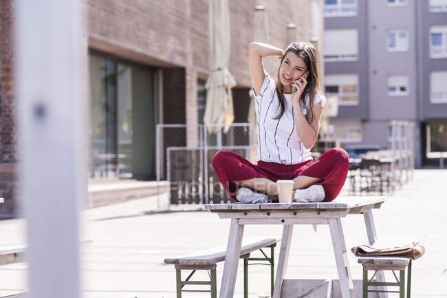 Молодая женщина, сидящая на столе в пивном саду и разговаривающая по телефону — стоковое фото