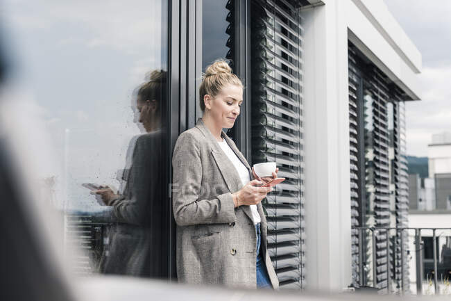 Femme d'affaires avec téléphone portable et tasse de café debout sur la terrasse du toit — Photo de stock