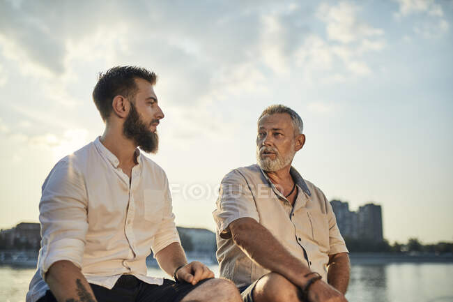 Отец и взрослый сын сидят на стене у реки и разговаривают. — стоковое фото