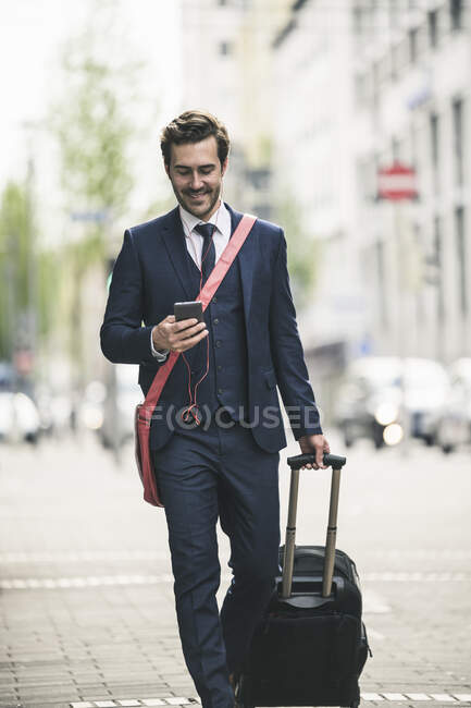 Hombre de negocios sonriente caminando por la ciudad con teléfono celular y maleta — Stock Photo