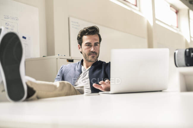Young businessman working relaxed in modern office, using laptop with feet on desk — Stock Photo