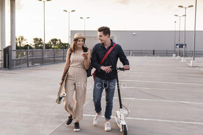 Happy young couple with cell phone, longboard and electric scooter on parking deck — Stock Photo
