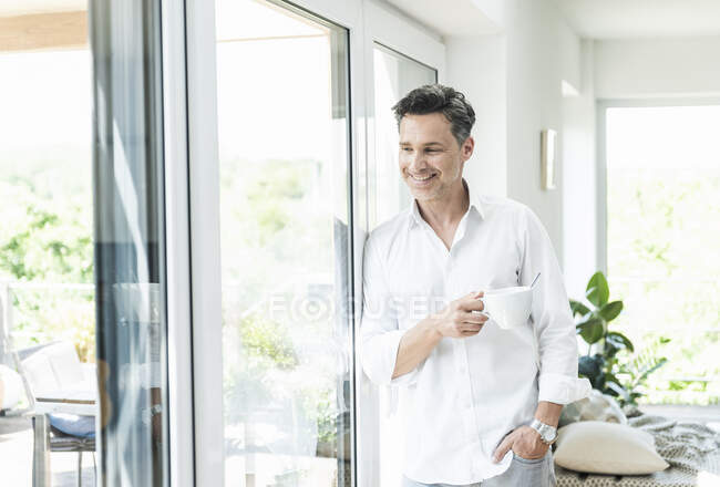 Uomo maturo appoggiato alla finestra, che beve caffè a casa — Foto stock