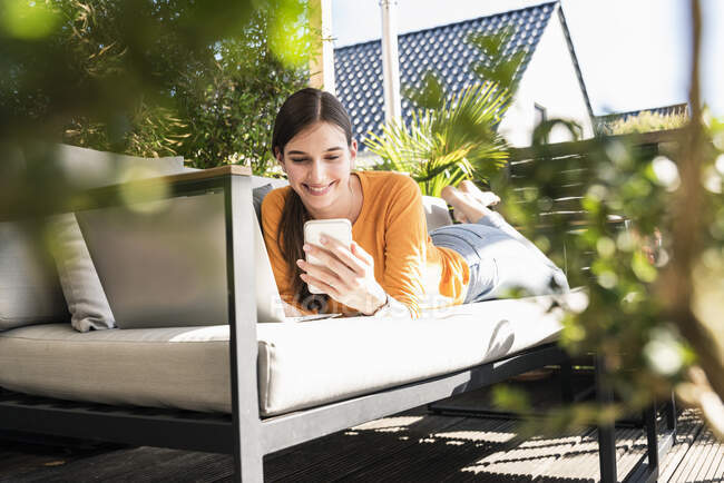 Mujer joven acostada en el sofá en la terraza utilizando el teléfono celular y el ordenador portátil — Stock Photo