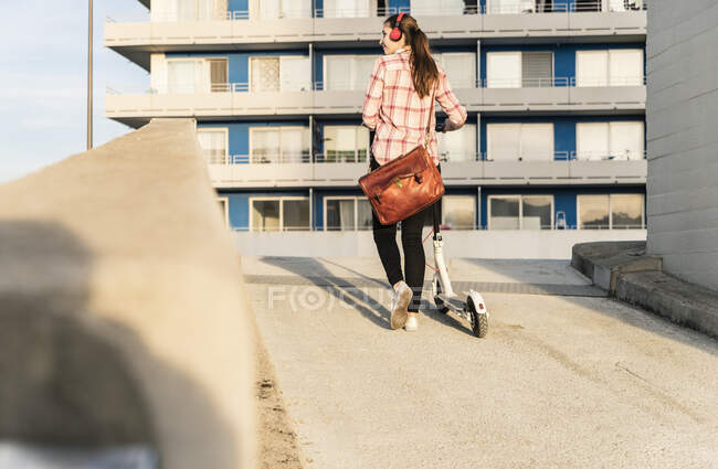 Giovane donna con cuffie spingendo scooter elettrico sul ponte di parcheggio — Foto stock