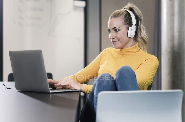 Femme d'affaires occasionnelle assise à la table au bureau en utilisant un ordinateur portable et en portant un casque — Photo de stock