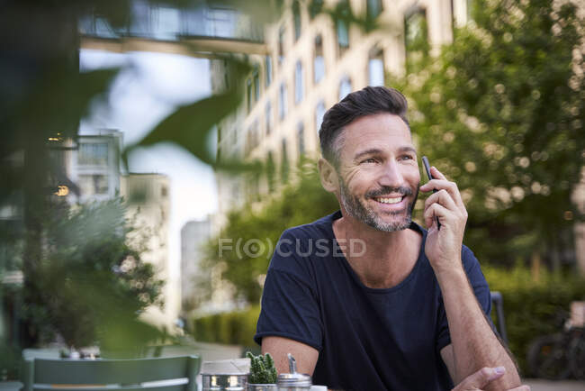 Homme mûr souriant utilisant un smartphone, assis au café de la rue — Photo de stock