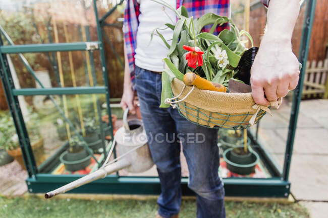 Nahaufnahme eines Mannes, der einen Korb mit Blumen und Gießkanne vor dem Gewächshaus im Garten hält — Stockfoto