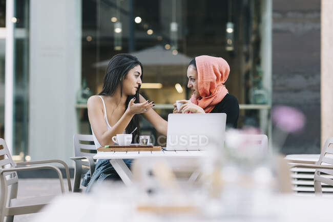 Two friends sitting together at a pavement cafe with laptop talking — Stock Photo