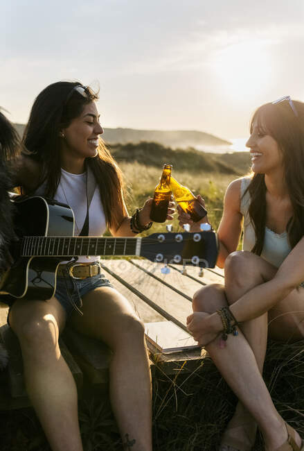 Duas mulheres felizes com guitarra clinking garrafas de cerveja no calçadão em dunas — Fotografia de Stock