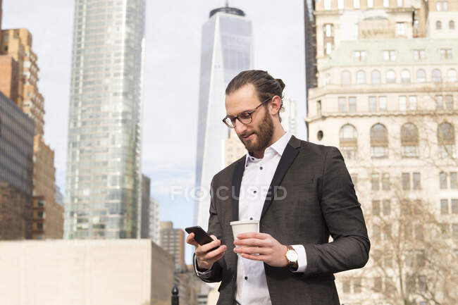 Empresario joven barbudo con café para ir a buscar en el teléfono celular, Nueva York, EE.UU. - foto de stock