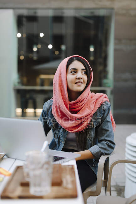 Усміхнена молода жінка з ноутбуком носить хустку в кафе на тротуарі — стокове фото