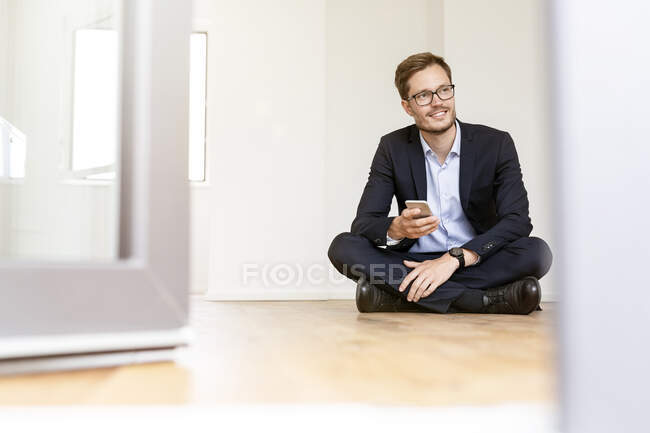 Hombre de negocios sonriente sentado en el suelo con teléfono celular - foto de stock