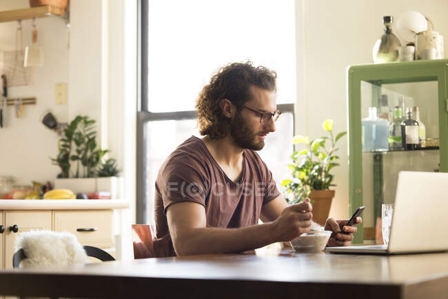 Людина сидить за столом з мискою муеслі дивлячись на смартфон. — стокове фото