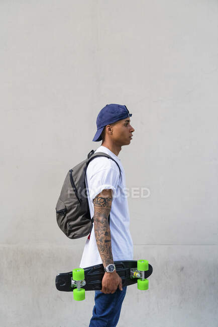 Jeune homme tatoué avec skateboard et sac à dos devant fond gris — Photo de stock