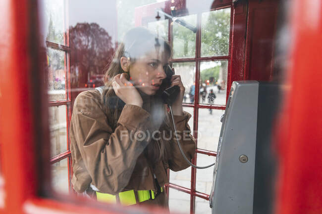 Mujeres jóvenes haciendo una llamada desde una cabina telefónica roja en la ciudad, Londres, Reino Unido - foto de stock