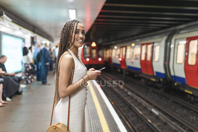 Portrait of smiling young woman with smartphone waiting at subway station platform, London, UK — Stock Photo