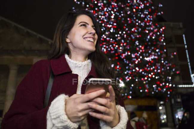 Portrait of happy young woman with smartphone in front of lighted Christmas tree outdoors — Stock Photo
