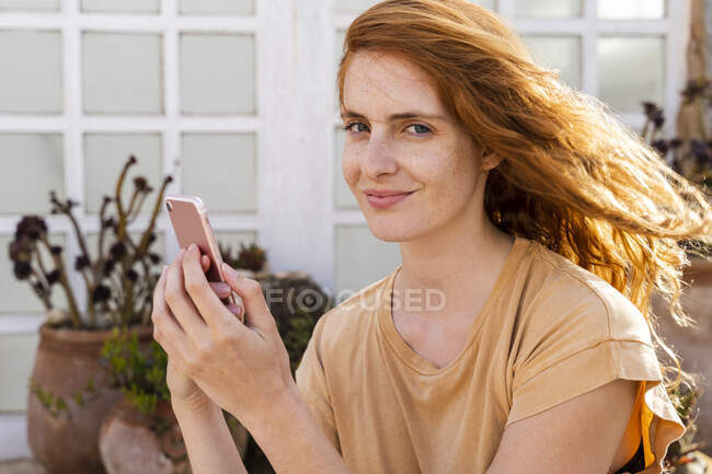 Retrato de una joven pelirroja sonriente con smartphone en la terraza - foto de stock