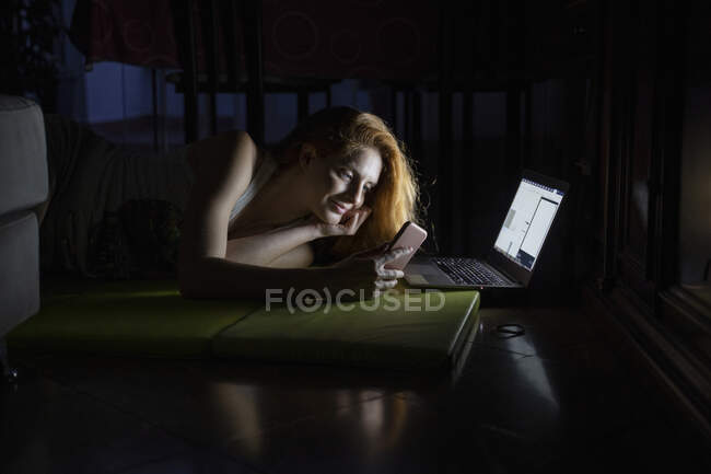 Mujer joven usando el ordenador portátil y el teléfono celular tarde en la noche - foto de stock