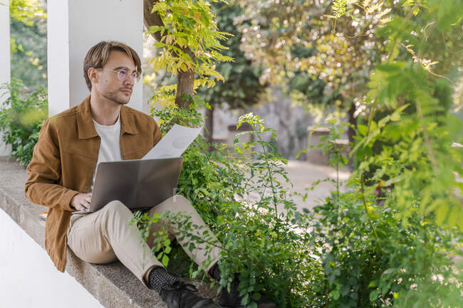 Porträt eines nachdenklichen Mannes, der auf einer Laubenwand sitzt und am Laptop arbeitet — Stockfoto