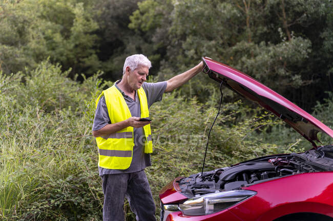 Senior man standing at his broken car wearing a safety vest and using his smartphone — Stock Photo